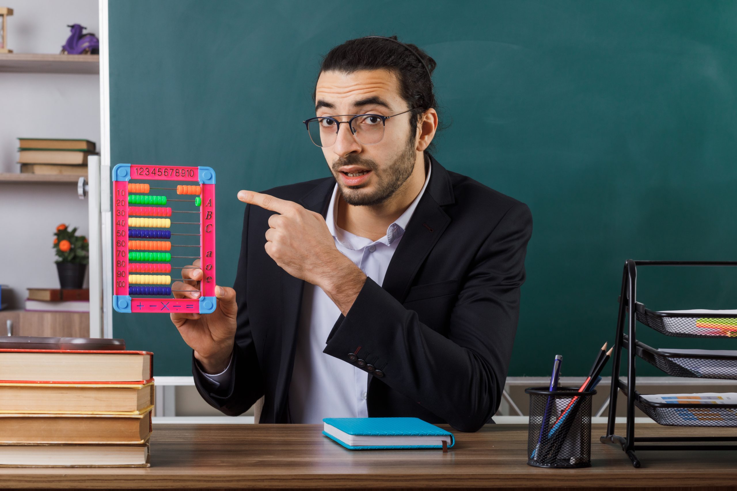 surprised-male-teacher-wearing-glasses-holding-points-abacus-sitting-table-with-school-tools-classroom
